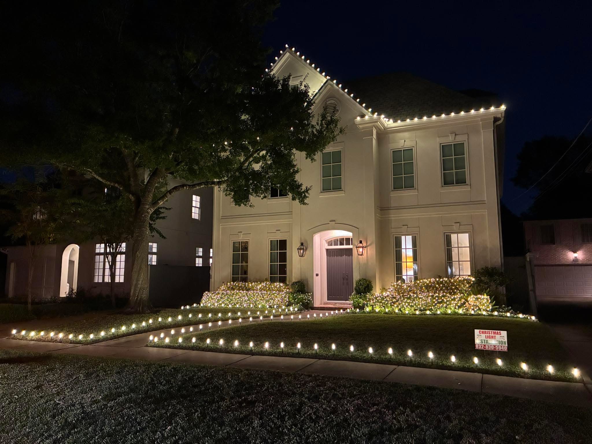 Elegant stucco home with warm white lights along the roofline, lit shrubs, and pathway lighting to the front door.