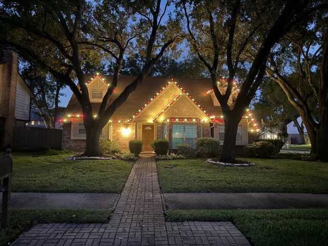 Brick home at dusk with warm and colored lights along the roof peaks, framed by two large trees.