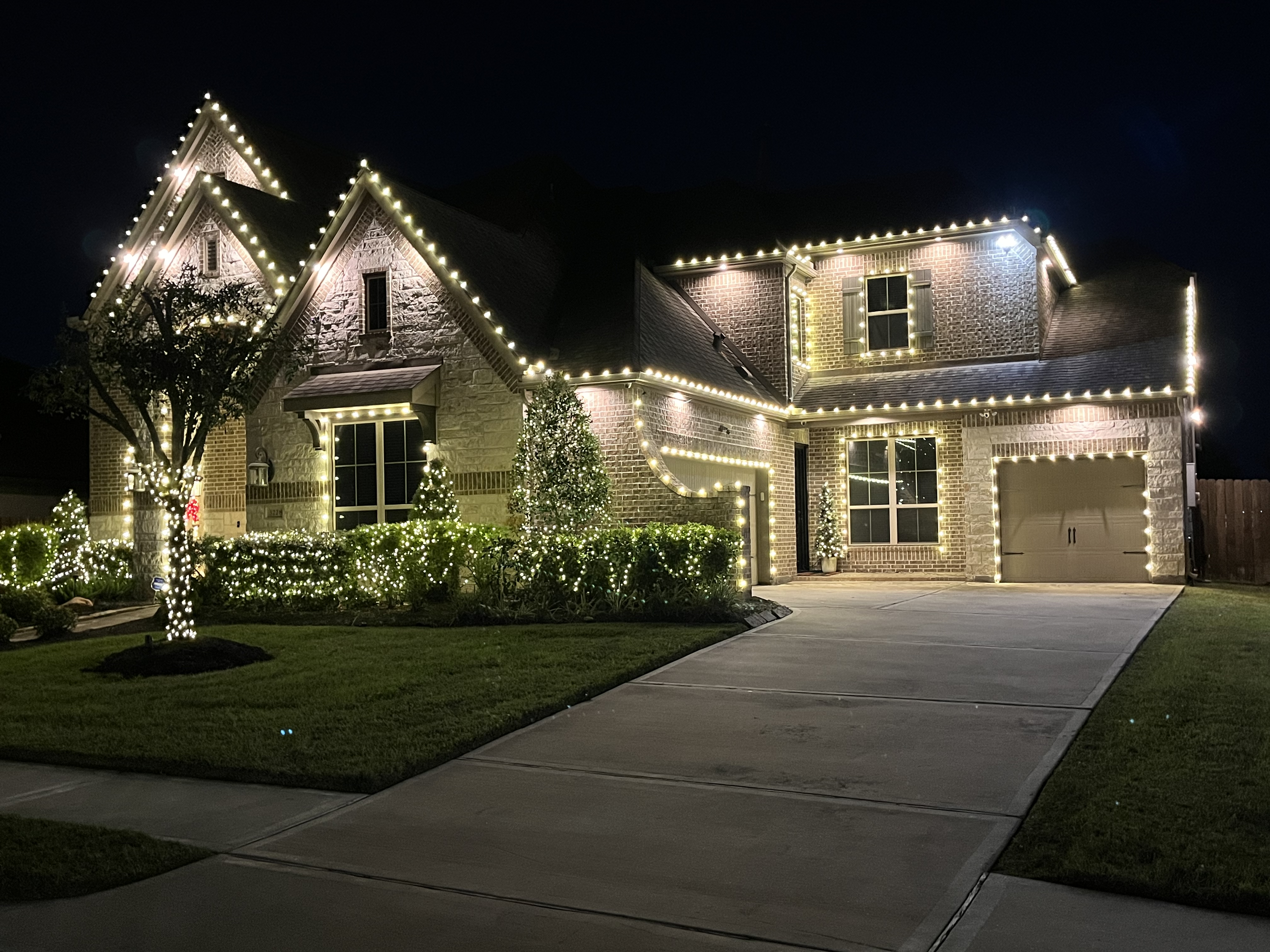 Large stone and brick home at night with warm white lights along every roofline and peak, garland-lit hedges, and a lit tree in the front yard.